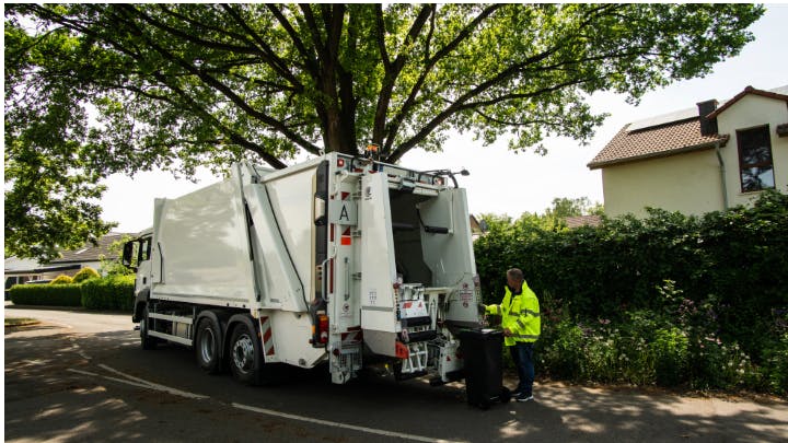 A waste disposal truck employee prepares to place an open bin on the truck's lifting apparatus. Cameras mounted on arms above will scan the interior of the bin for contaminants before the bin is dumped. If contaminants are detected, the bin will be returned to the customer unemptied.