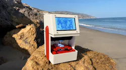 White-and-red SCANIMUS research-use-only scanning-microscopy device positioned on a sunlit coastal rock, with a sandy beach, ocean, cliffs, and blue sky behind it.