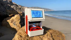 White-and-red SCANIMUS research-use-only scanning-microscopy device positioned on a sunlit coastal rock, with a sandy beach, ocean, cliffs, and blue sky behind it.